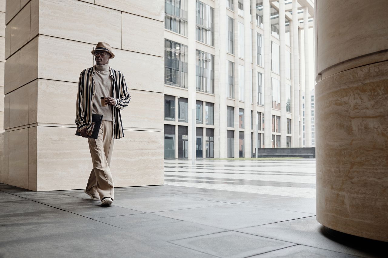 Fashionable man wearing hat and striped jacket walking in a modern cityscape.