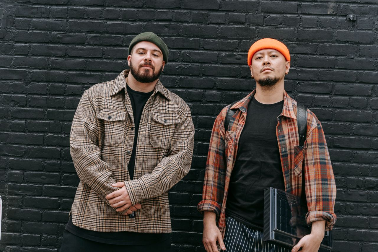 Two men in fashionable checkered jackets pose against a black brick wall outdoors.