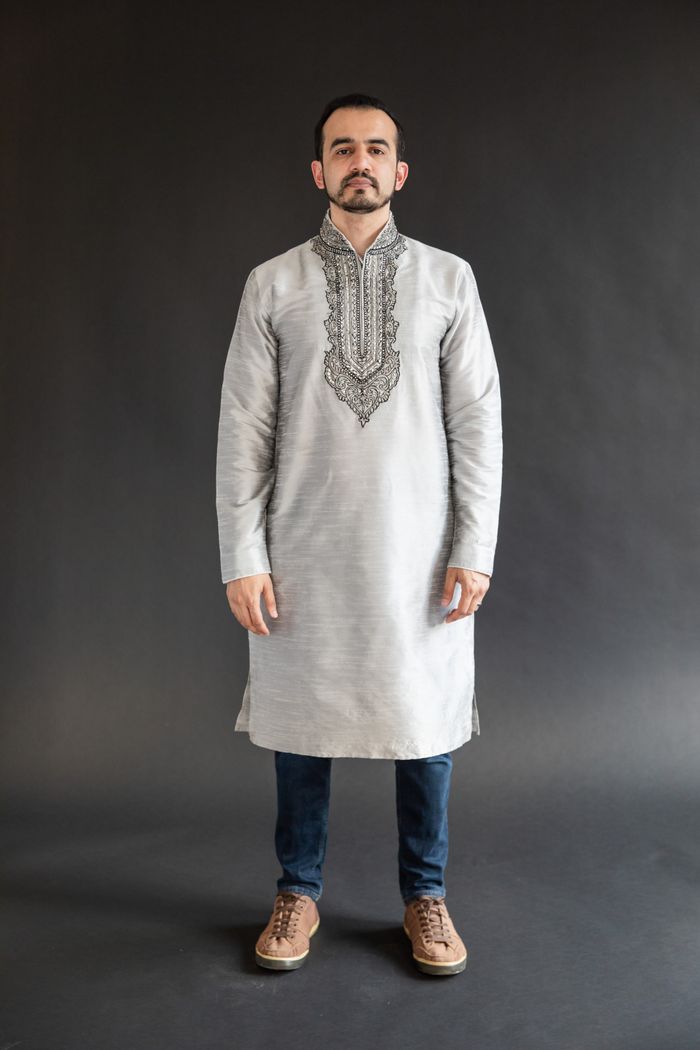 Portrait of a man wearing a traditional embroidered kurta standing against a dark studio backdrop.